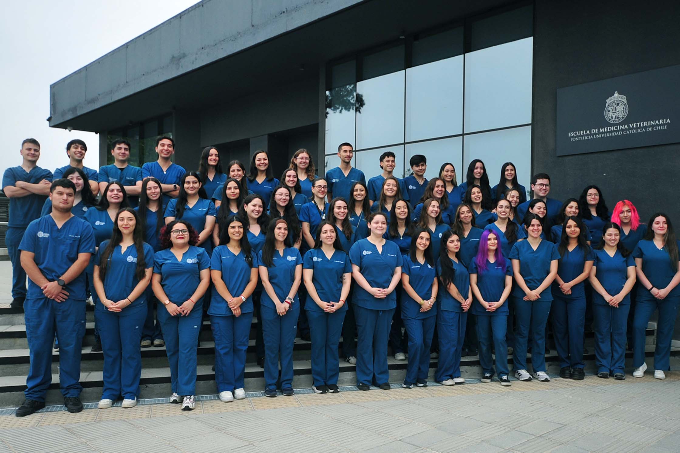 Alumni de carrera Medicina Veterinaria UC posando con traje clínico frente a la Escuela de Medicina Veterinaria.
